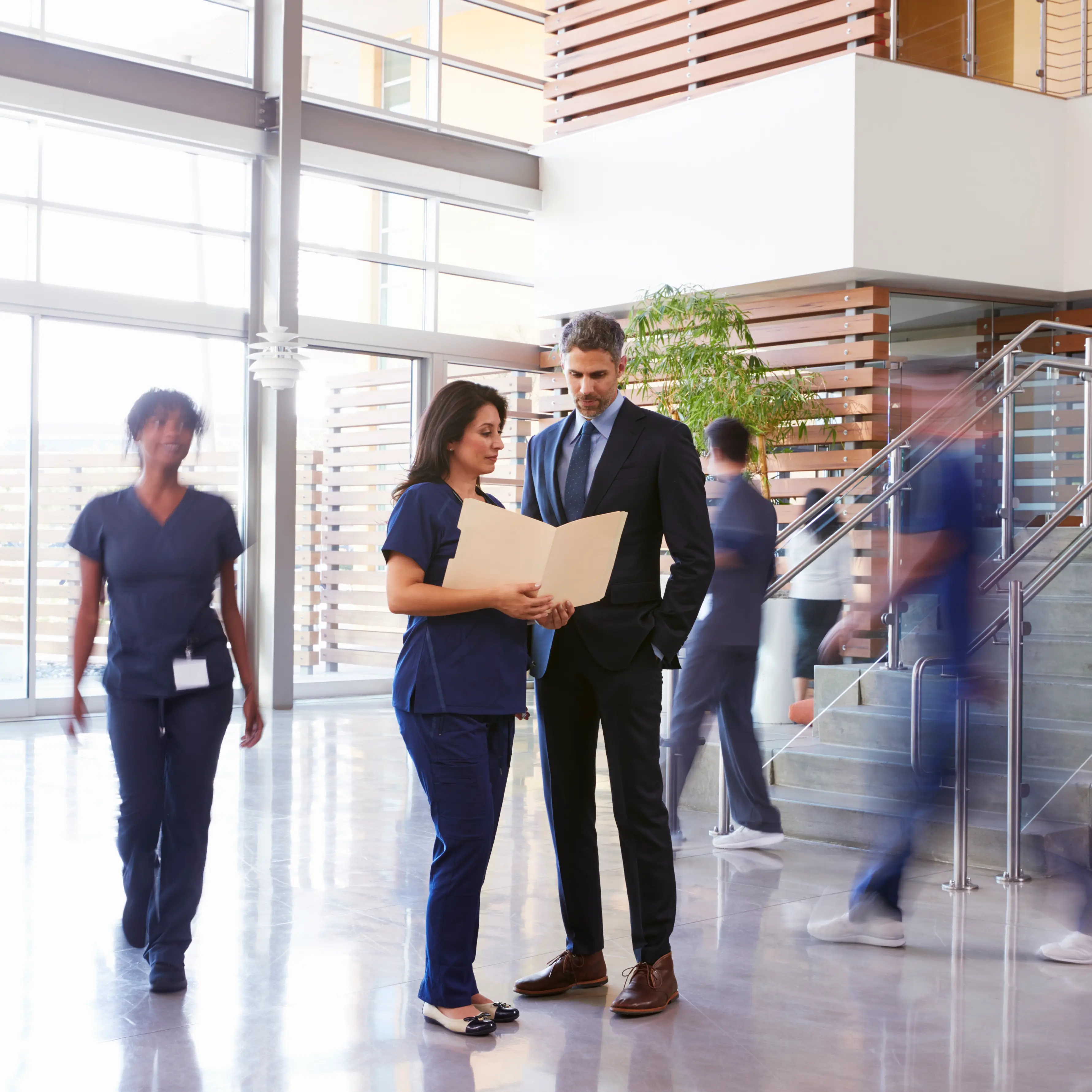 A female healthcare professional in blue scrubs and a male executive in a dark suit reviewing documents in a folder. They are standing in a bright, modern hospital lobby with high ceilings, large windows, and a staircase. Other medical staff are seen walking in the background, representing a busy clinical environment.
