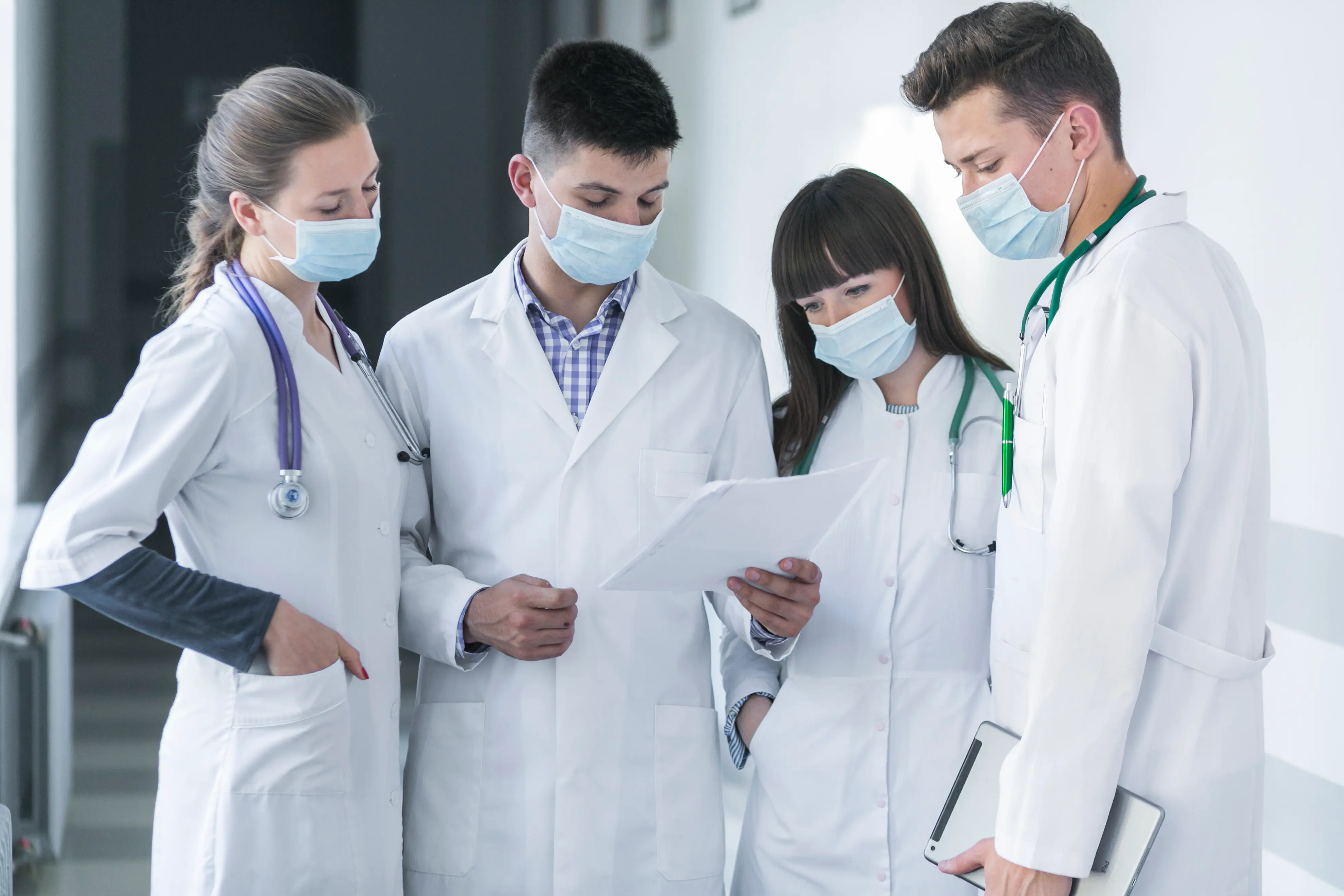 A group of four diverse healthcare professionals in white lab coats and surgical masks standing in a bright hospital corridor. They are huddled together looking at a document held by one of the male doctors, representing a collaborative interdisciplinary medical team